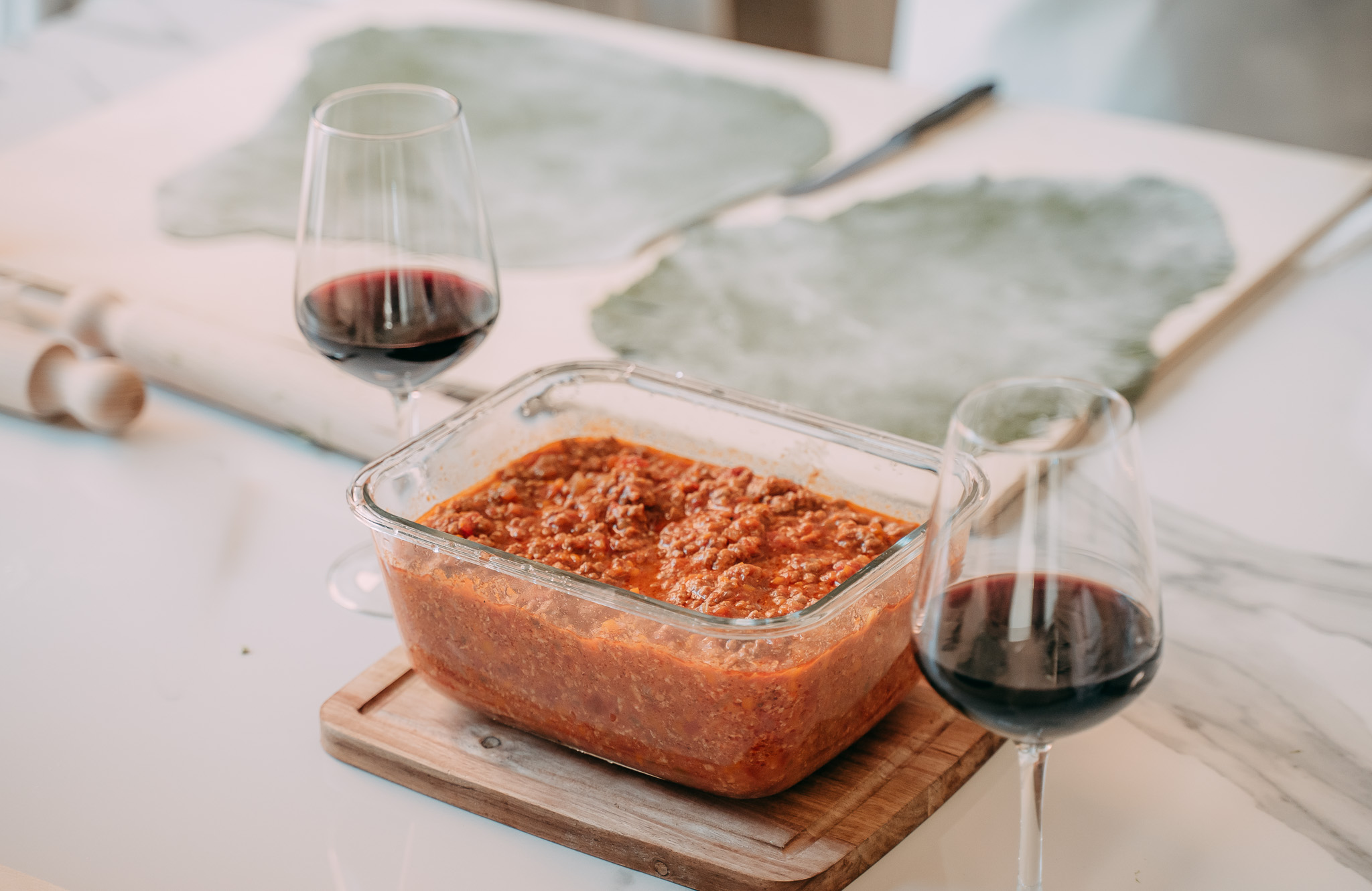 Contenitore con ragù di carne su tagliere in legno accanto a due bicchieri di vino rosso