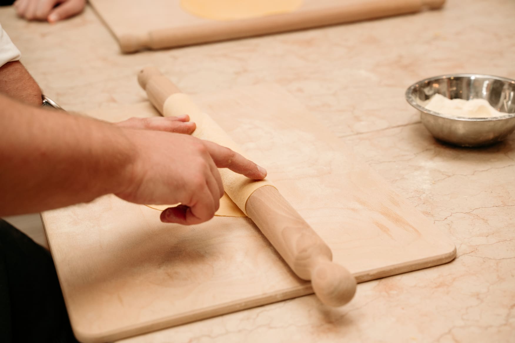Close-up of a person's hands using a wooden rolling pin to wrap a sheet of fresh pasta dough on a floured wooden board, during a pasta making class