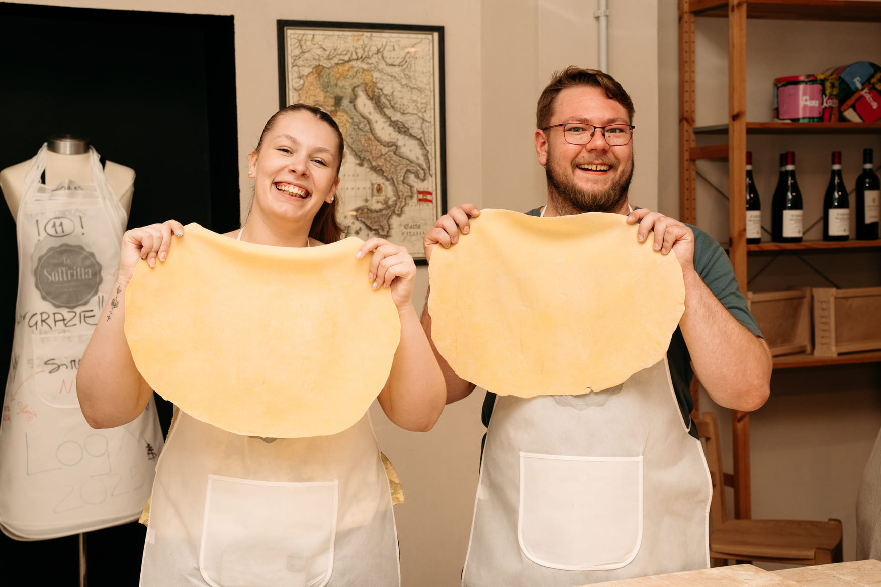 A woman and a man proudly holding up large, flat sheets of freshly made pasta dough