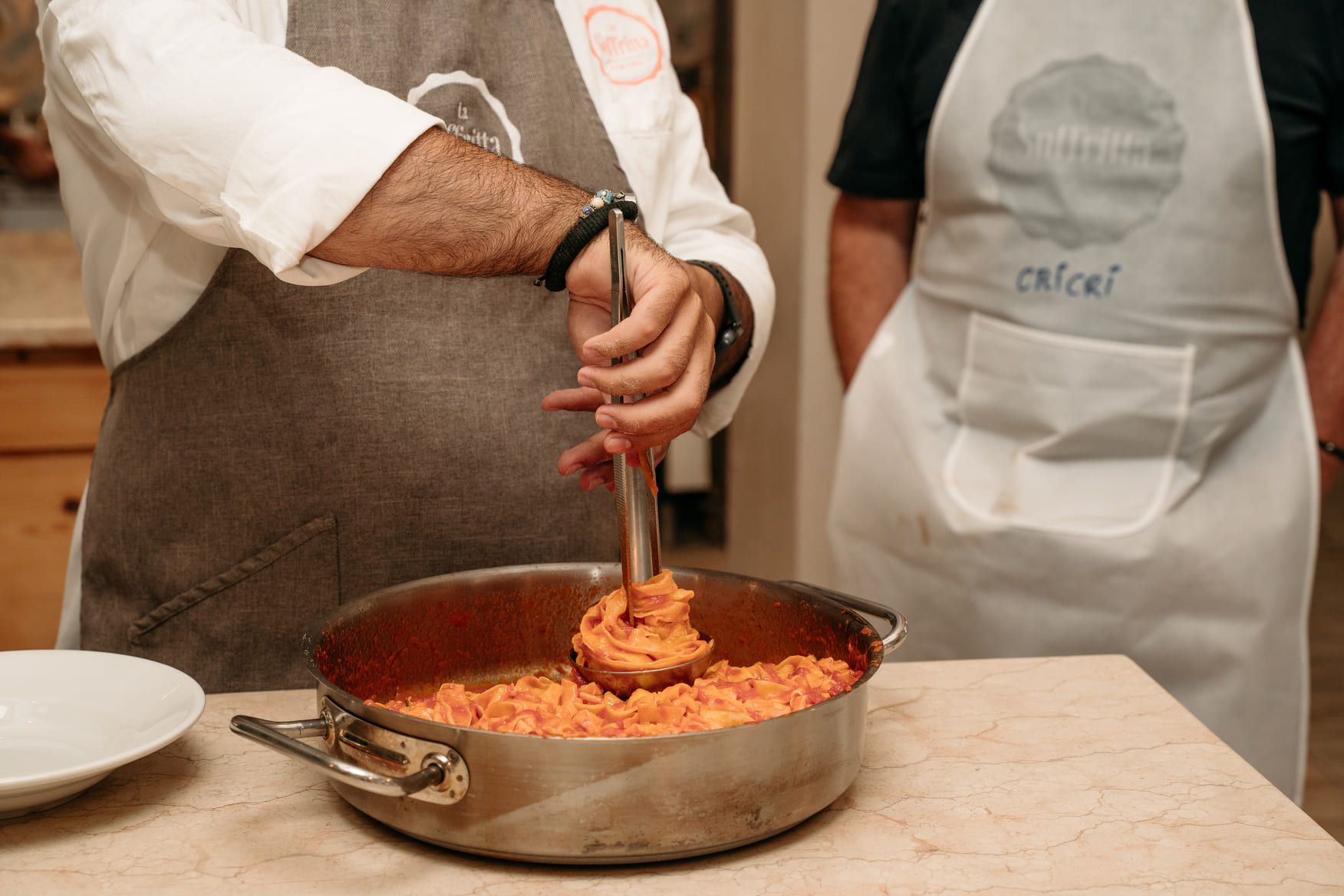 Chef stirs a large, stainless steel pot of bubbling pasta sauce with a metal ladle, showing a cooking class in action